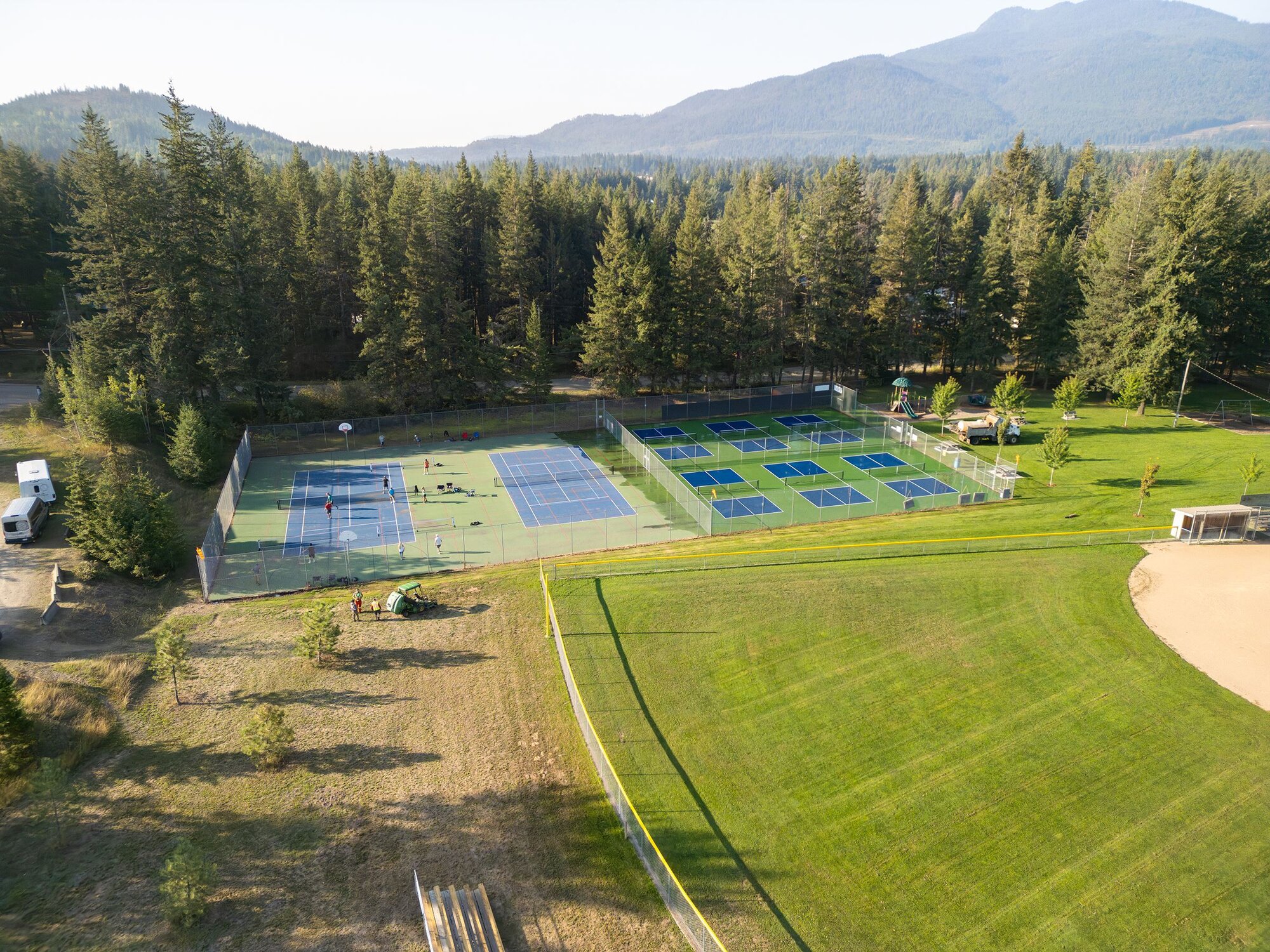 An aerial view of the tennis courts, ball field, and green space at Klahani Park, framed by forest and mountains